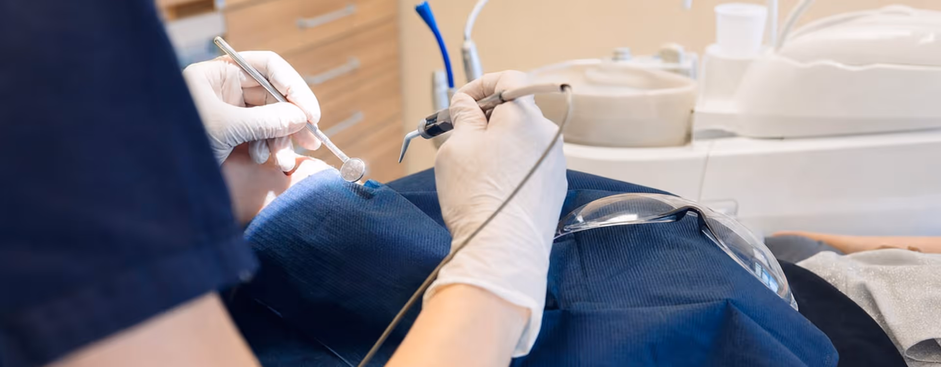 Zahnmedizinische Fachkraft in dunkelblauer Kleidung und weißen Handschuhen behandelt einen Patienten mit Spiegel und Instrument im Behandlungszimmer.